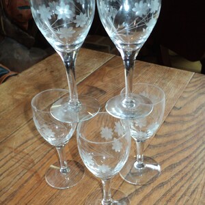 May include: Five clear wine glasses with etched floral designs. Two taller glasses are balanced on top of three smaller glasses. The glasses are arranged on a wooden surface, showcasing the delicate floral patterns.
