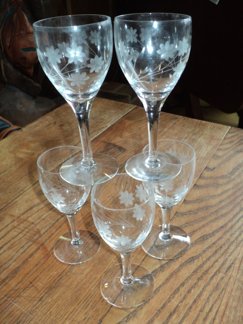 May include: Five clear wine glasses with etched floral designs. Two taller glasses are balanced on top of three smaller glasses. The glasses are arranged on a wooden surface, showcasing the delicate floral patterns.