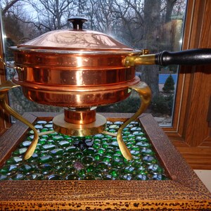May include: A copper chafing dish with a black handle and a brass stand. The stand is on a wooden base with green glass beads.