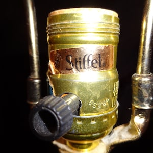 May include: Close-up of a brass lamp socket with a black knob and a label that reads "Stiffel." The socket is attached to a gold-colored metal frame. The background is black.