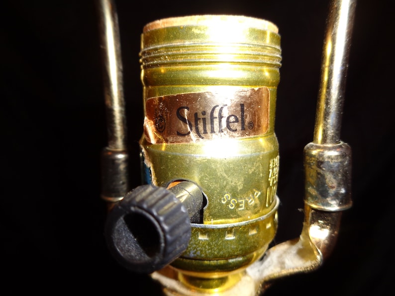 May include: Close-up of a brass lamp socket with a black knob and a label that reads "Stiffel." The socket is attached to a gold-colored metal frame. The background is black.