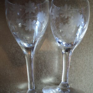 May include: Two clear wine glasses with etched floral designs. The glasses have a slender stem and a wide, round base. The floral pattern is repeated on both glasses, creating a matching set. The glasses are made of clear glass.