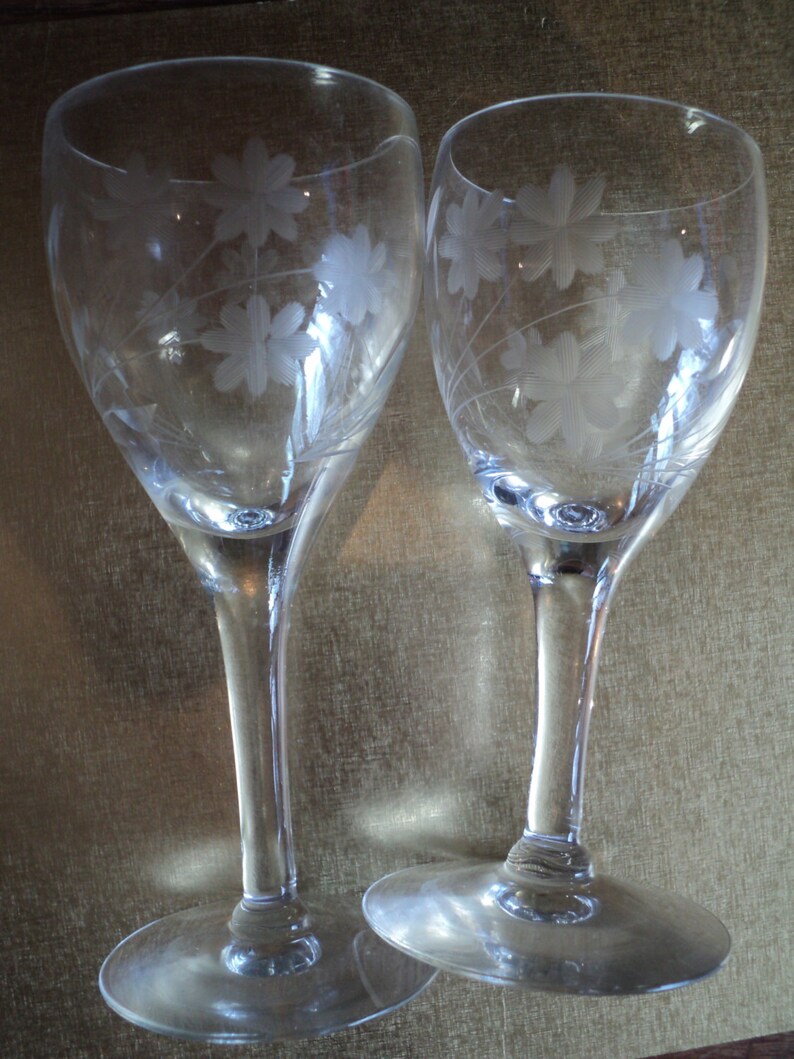 May include: Two clear wine glasses with etched floral designs. The glasses have a slender stem and a wide, round base. The floral pattern is repeated on both glasses, creating a matching set. The glasses are made of clear glass.