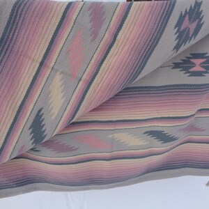 May include: A woven throw blanket with a geometric pattern in shades of pink, gray, and beige. The blanket is folded in half and has a fringe on the bottom edge.