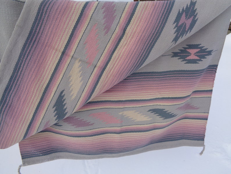 May include: A woven throw blanket with a geometric pattern in shades of pink, gray, and beige. The blanket is folded in half and has a fringe on the bottom edge.