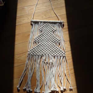 May include: A cream-colored macrame wall hanging with a tree design. The handmade piece features a woven pattern forming a tree shape, with long, fringed strands hanging below. The wall decor is suspended from a wooden dowel and a hanging cord.