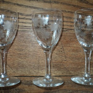 May include: Set of three clear glass wine glasses with etched floral designs. The glasses have a slender stem and a round base. The etched design features a cluster of flowers and leaves. The glasses are displayed on a wooden surface.