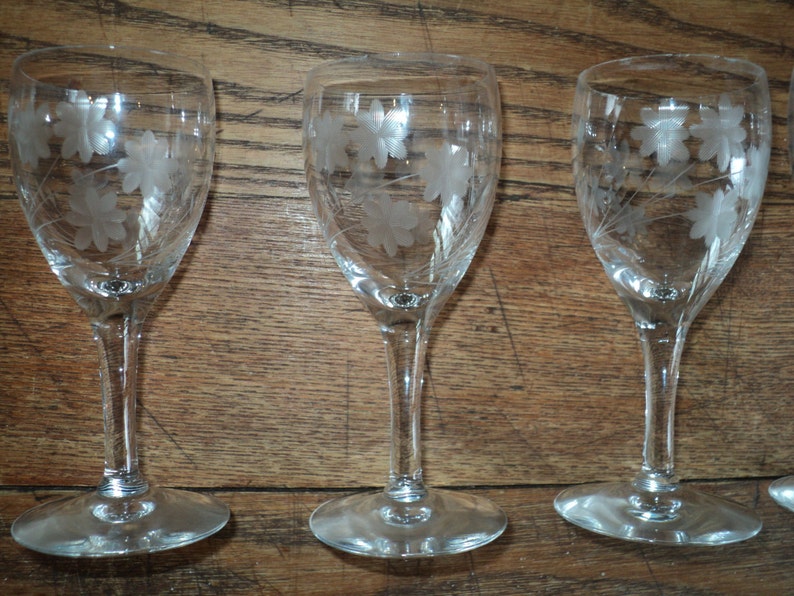 May include: Set of three clear glass wine glasses with etched floral designs. The glasses have a slender stem and a round base. The etched design features a cluster of flowers and leaves. The glasses are displayed on a wooden surface.