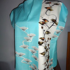 May include: A vintage turquoise blouse with a white panel featuring a botanical print. The blouse has a unique design with short sleeves and a draped neckline. The botanical print includes fan-like shapes and branches with leaves.