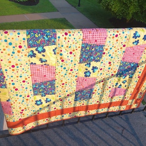 May include: A colorful patchwork quilt with a yellow background covered in polka dots. The quilt features squares of pink, blue, and floral patterns, with an orange border. The quilt is draped over a black railing.