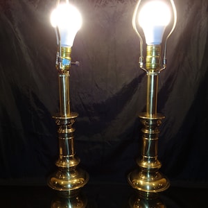 May include: Two brass table lamps with a classic design. Each lamp features a bulb, a decorative base, and a tall, slender stem. The lamps are lit, emitting a warm glow against a dark background. The lamps are a matching set.