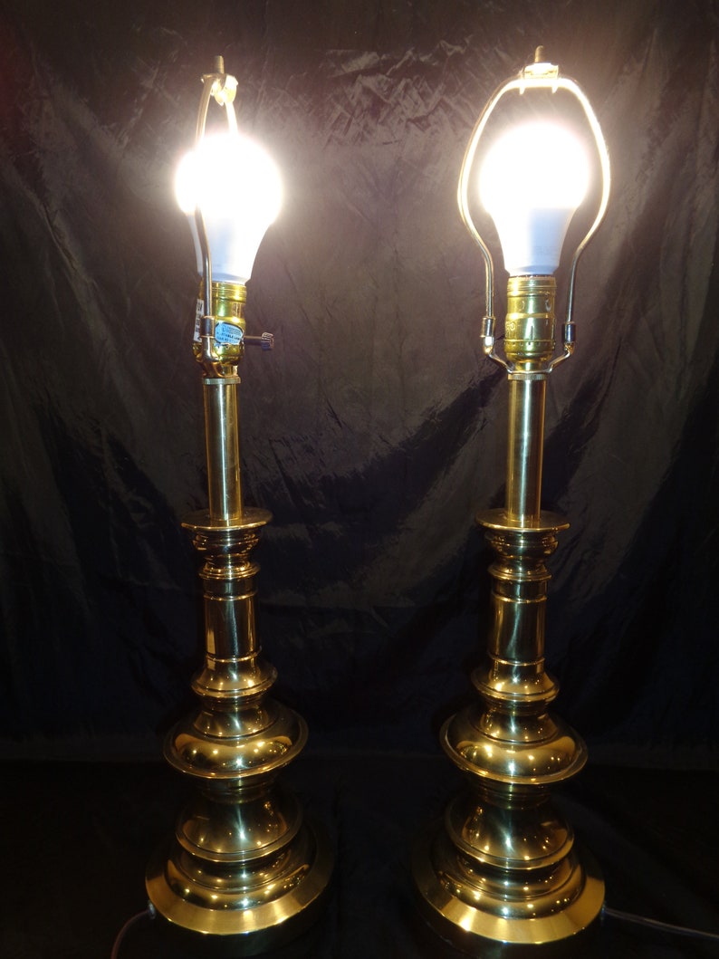 May include: Two brass table lamps with a classic design. Each lamp features a bulb, a decorative base, and a tall, slender stem. The lamps are lit, emitting a warm glow against a dark background. The lamps are a matching set.
