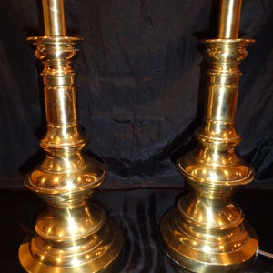 May include: Two vintage brass table lamps. Each lamp has a detailed, ornate base and a tall, slender stem. The lamps are a bright gold color and are set against a dark background. The lamps are plugged in and ready to use.