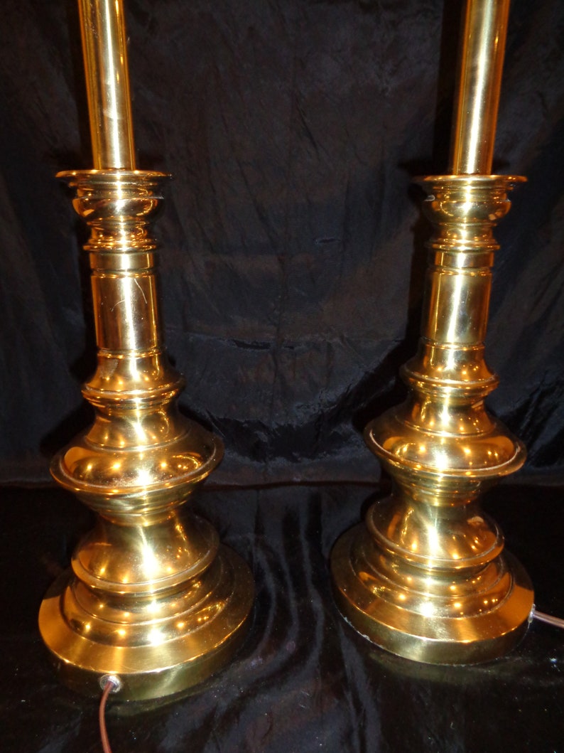 May include: Two vintage brass table lamps. Each lamp has a detailed, ornate base and a tall, slender stem. The lamps are a bright gold color and are set against a dark background. The lamps are plugged in and ready to use.