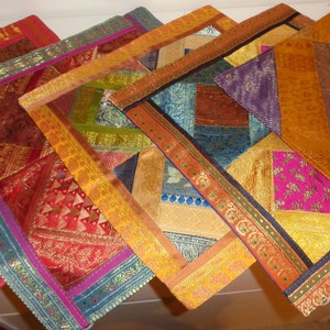 May include: Five colorful square throw pillows with intricate patterns and gold trim. The pillows are made of a variety of fabrics, including silk and velvet. The pillows are arranged in a fan shape on a white surface.