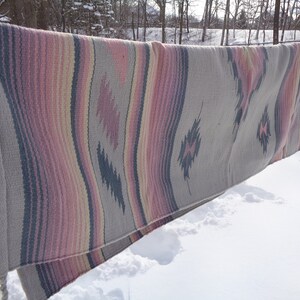 May include: A woven blanket with a Southwestern design in shades of pink, gray, and white. The blanket is hanging on a clothesline outside in the snow.