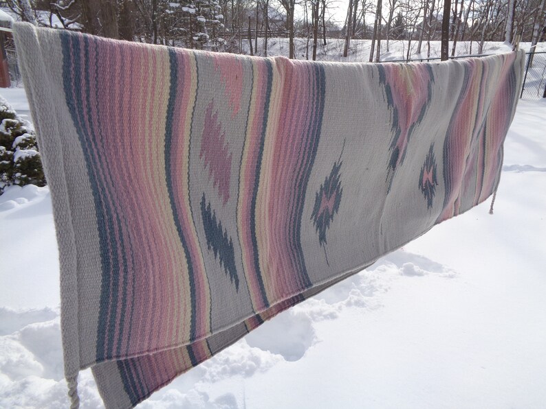 May include: A woven blanket with a Southwestern design in shades of pink, gray, and white. The blanket is hanging on a clothesline outside in the snow.