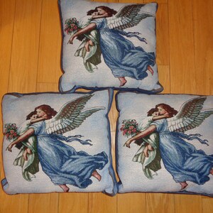 Op de afbeelding: Drie vierkante decoratieve kussens met een tapijtontwerp. Elk kussen toont een engel in een vloeiende blauwe jurk, die een kind vasthoudt. De engel heeft grote, gedetailleerde vleugels en de kussens hebben een blauwe rand.