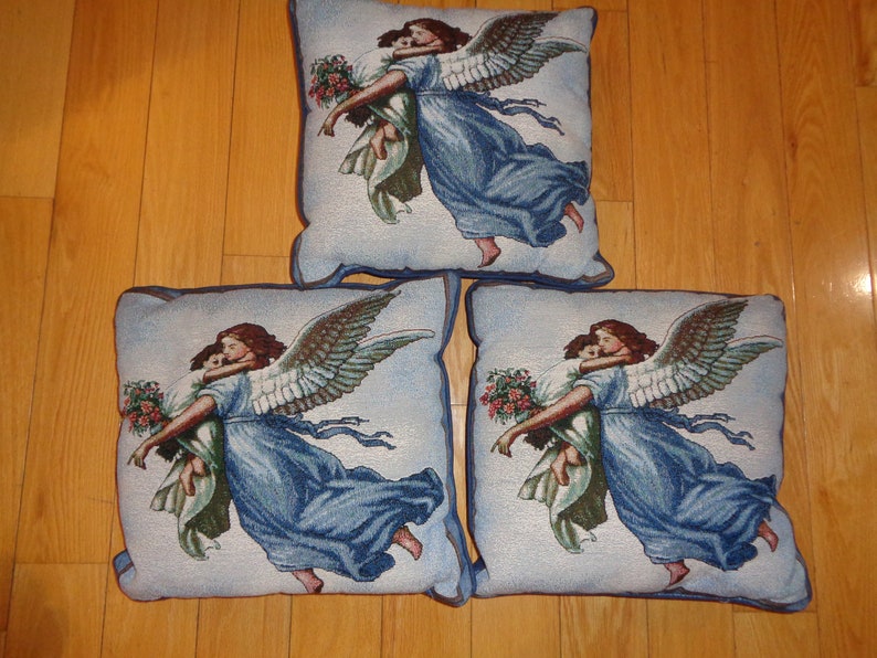 Op de afbeelding: Drie vierkante decoratieve kussens met een tapijtontwerp. Elk kussen toont een engel in een vloeiende blauwe jurk, die een kind vasthoudt. De engel heeft grote, gedetailleerde vleugels en de kussens hebben een blauwe rand.