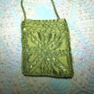 May include: A small, olive green beaded evening bag with a long, beaded strap. The bag is rectangular with a geometric beaded design on the front. The background is a white crocheted doily on a light blue surface.