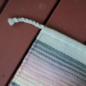 May include: A woven rug with a fringe edge. The rug has stripes of pink, light green, gray, and white.