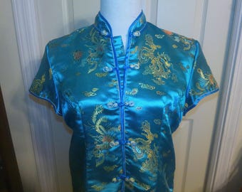 May include: A teal-colored, short-sleeved top with a mandarin collar and frog closures. The top is adorned with gold embroidered dragons and cloud motifs. The edges are trimmed with a darker teal piping. The fabric has a satin sheen.