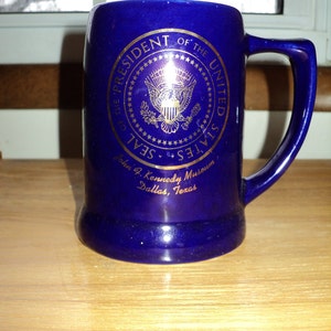 May include: A blue ceramic mug with a gold Presidential Seal of the United States. The mug reads "President of the United States Seal John F. Kennedy Museum Dallas, Texas".