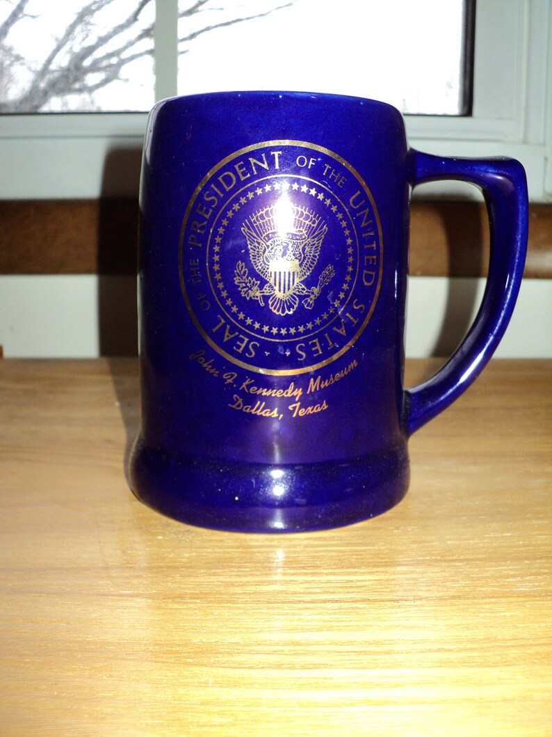 May include: A blue ceramic mug with a gold Presidential Seal of the United States. The mug reads "President of the United States Seal John F. Kennedy Museum Dallas, Texas".