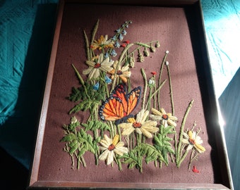 May include: Embroidered wall art featuring a butterfly with orange, black, and white wings surrounded by yellow and white flowers on a brown background.