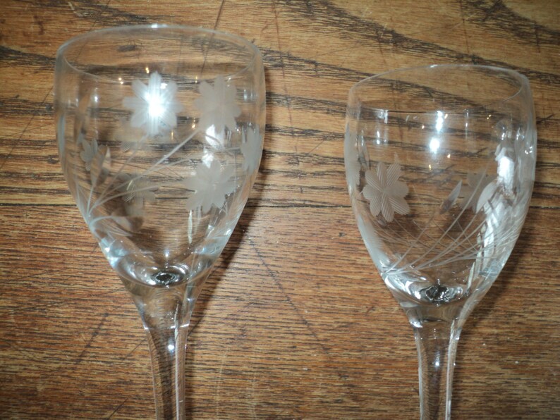 May include: Two clear wine glasses with etched floral designs. The glasses have long stems and a delicate, elegant appearance. The floral pattern includes stylized flowers and leaves, adding a touch of sophistication to the glassware.