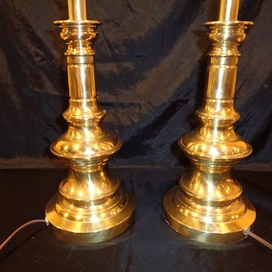 May include: Two vintage brass table lamps. Each lamp has a tiered base, a central column with decorative molding, and a top section for the light fixture. The lamps are made of polished brass and have a warm, golden color.