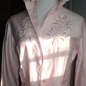 Pu&ograve; includere: Camicia rosa chiaro con bottoni, colletto ampio e maniche a tre quarti. La camicia presenta ricami floreali con dettagli di paillettes sul petto e sul colletto. La camicia &egrave; esposta su un manichino.