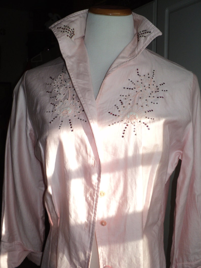 Pu&ograve; includere: Camicia rosa chiaro con bottoni, colletto ampio e maniche a tre quarti. La camicia presenta ricami floreali con dettagli di paillettes sul petto e sul colletto. La camicia &egrave; esposta su un manichino.