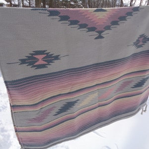 May include: A woven throw blanket with a geometric pattern in shades of pink, gray, and black. The blanket has a fringe border.