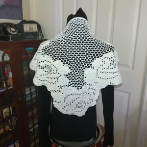 May include: A white, crocheted shawl with a floral design. The shawl is draped over a black top, showcasing the intricate openwork and the detailed rose pattern. The shawl's edges have a scalloped finish.