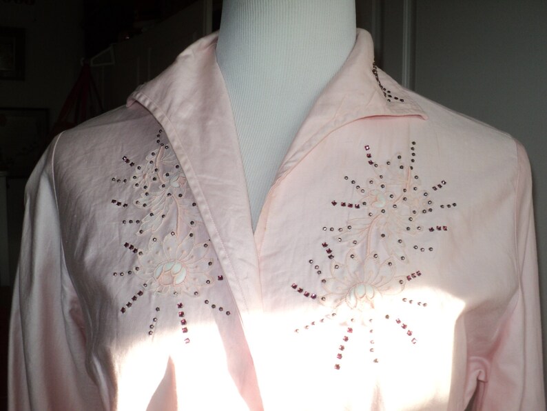 Pu&ograve; includere: Camicia rosa pallido con colletto e maniche lunghe. La camicia presenta ricami floreali con piccoli abbellimenti rosso scuro. La camicia &egrave; esposta su un manichino.