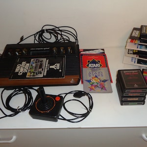 May include: A vintage Atari Video Computer System with two joysticks, a game cartridge, and a stack of game cartridges. The system is brown and black with a woodgrain finish. The cartridges are black with colorful labels.