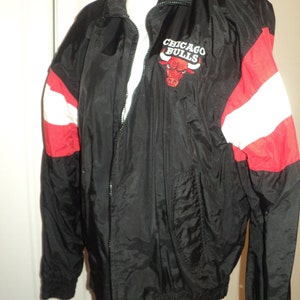 K&ouml;nnte beinhalten: Schwarze Windbreaker-Jacke mit roten und wei&szlig;en Streifen an den &Auml;rmeln. Die Jacke hat einen Rei&szlig;verschluss und ein Logo mit der Aufschrift "Chicago Bulls" und einem roten Bullen als Maskottchen.