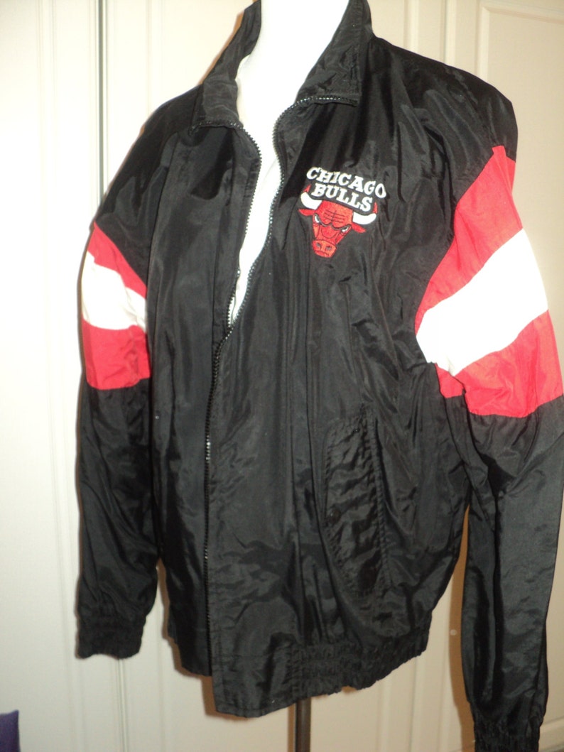 K&ouml;nnte beinhalten: Schwarze Windbreaker-Jacke mit roten und wei&szlig;en Streifen an den &Auml;rmeln. Die Jacke hat einen Rei&szlig;verschluss und ein Logo mit der Aufschrift "Chicago Bulls" und einem roten Bullen als Maskottchen.
