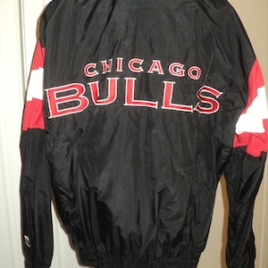 K&ouml;nnte beinhalten: Schwarze Windbreaker-Jacke mit roten und wei&szlig;en Akzenten. Auf der R&uuml;ckseite der Jacke befindet sich der gestickte Text "CHICAGO BULLS".
