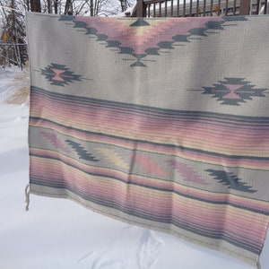 May include: A gray and pink woven throw blanket with a geometric pattern. The blanket is hanging on a clothesline outside in the snow.