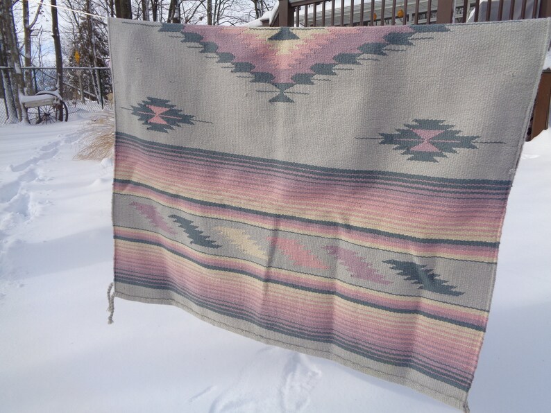May include: A gray and pink woven throw blanket with a geometric pattern. The blanket is hanging on a clothesline outside in the snow.