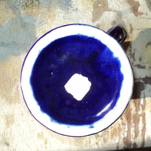 May include: A blue ceramic mug with a white rim and a white square in the center. The mug is sitting on a textured surface.