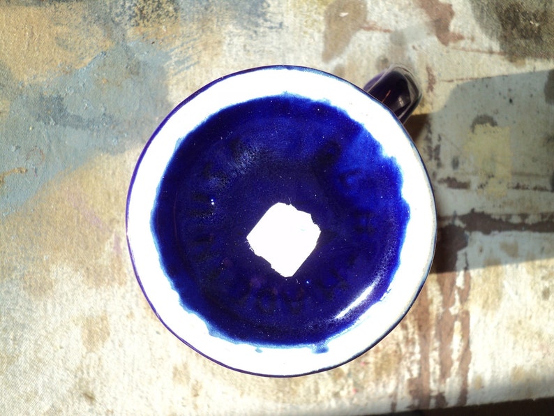 May include: A blue ceramic mug with a white rim and a white square in the center. The mug is sitting on a textured surface.