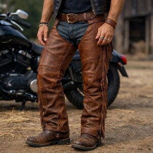 May include: Brown leather motorcycle chaps with fringe detailing and a zipper closure. The chaps are worn with a brown leather belt and boots. A black motorcycle is in the background.