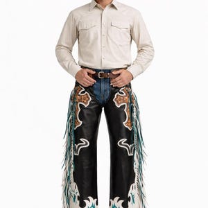May include: A person wearing a brown cowboy hat, light beige button-down shirt, and black leather chaps. The chaps feature decorative cross and floral designs with fringe and turquoise accents. The person is wearing brown cowboy boots.