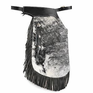 May include: A single leather chaps with a black and white cowhide pattern. The chaps have a black leather belt and fringe along the bottom edge. Silver-toned decorative accents are visible on the side.