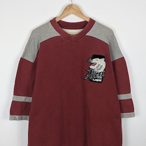 K&ouml;nnte beinhalten: Burgunderfarbenes V-Ausschnitt-Sweatshirt mit grauen &Auml;rmeln und Streifen. Mit einem Aufn&auml;her mit einem Tiger-Motiv und dem Text "Airex USA". Das Sweatshirt h&auml;ngt an einem Holzb&uuml;gel.
