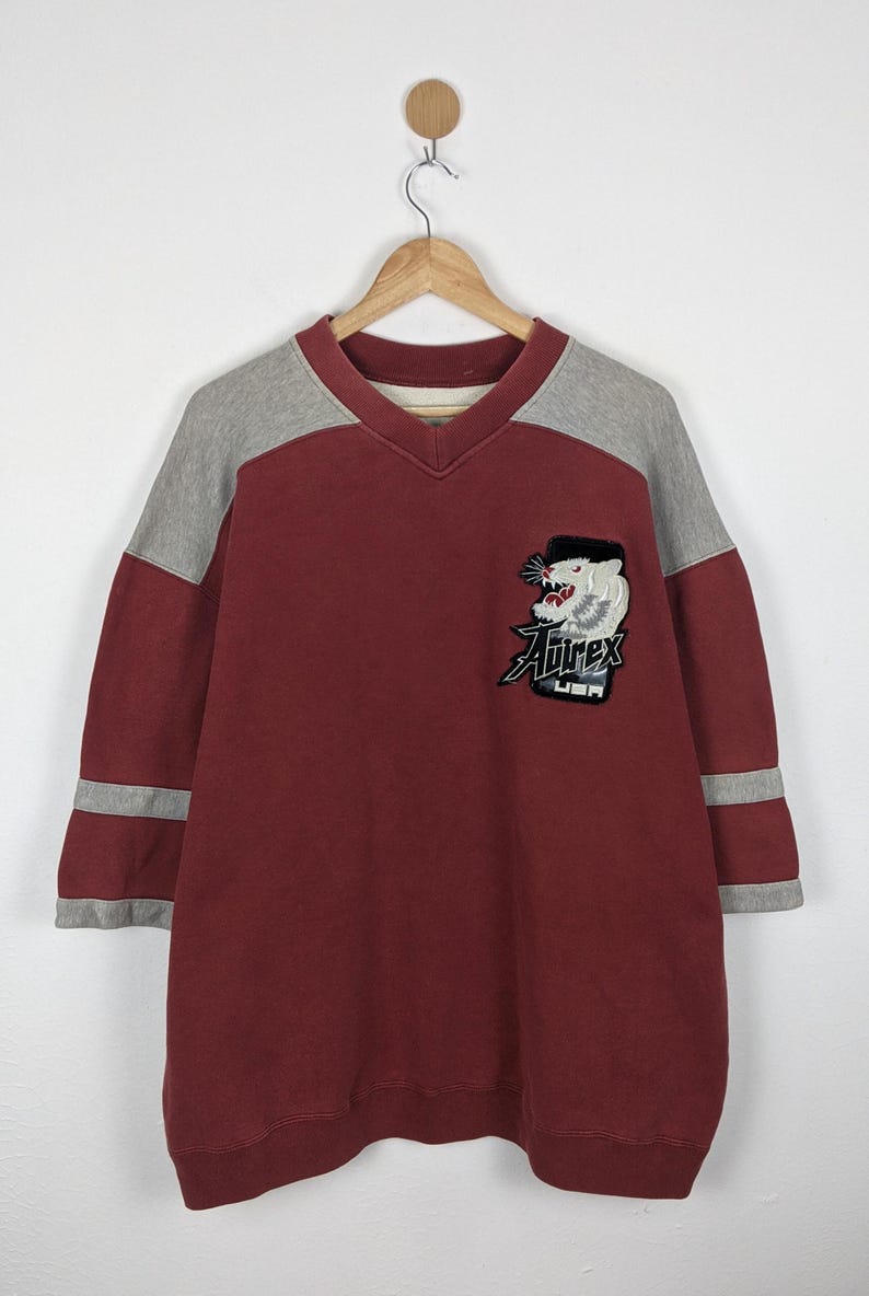 K&ouml;nnte beinhalten: Burgunderfarbenes V-Ausschnitt-Sweatshirt mit grauen &Auml;rmeln und Streifen. Mit einem Aufn&auml;her mit einem Tiger-Motiv und dem Text "Airex USA". Das Sweatshirt h&auml;ngt an einem Holzb&uuml;gel.