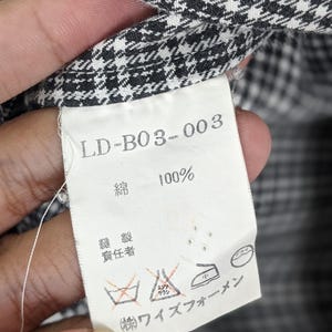 Puede incluir: Primer plano de una etiqueta de tela con el texto "LD-B03-003" y "100%" impreso. La etiqueta es blanca con texto negro y est&aacute; adherida a una tela con estampado de pata de gallo en blanco y negro. La etiqueta tambi&eacute;n incluye s&iacute;mbolos de cuidado.